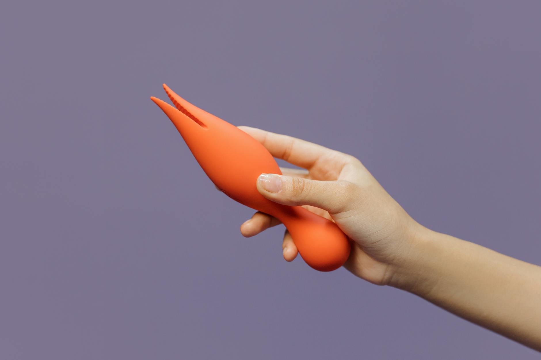 A hand holding a bright orange clitoral vibrator against a purple backdrop, symbolizing taking control of your own pleasure.