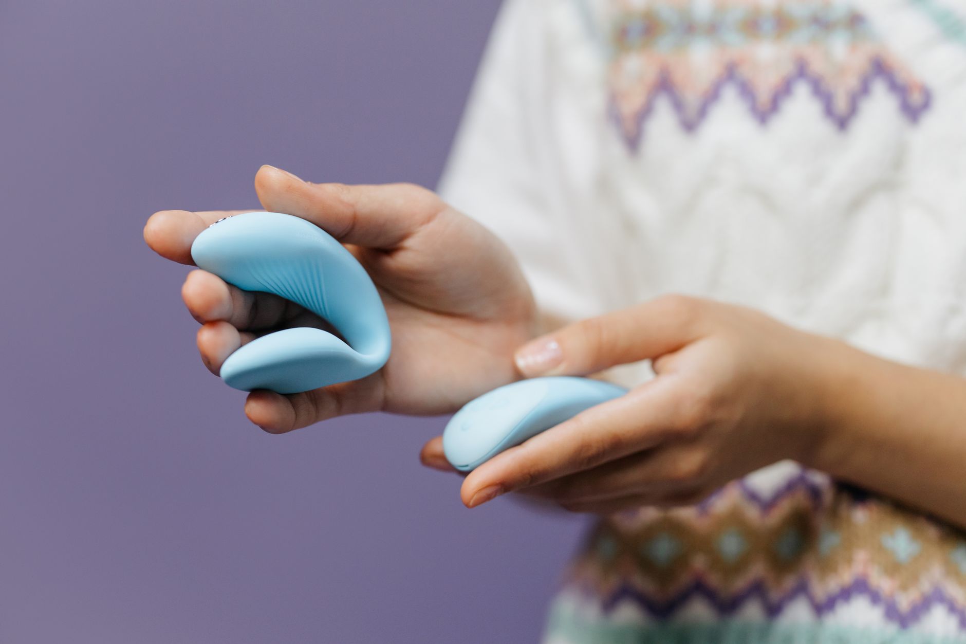 Close-up of hands holding a sleek blue clitoral vibrator against a purple background.